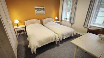 Una habitación con dos camas individuales, una mesita pequeña, una silla, una mesita de noche con lámpara, una ventana con cortinas y un cuadro en la pared.