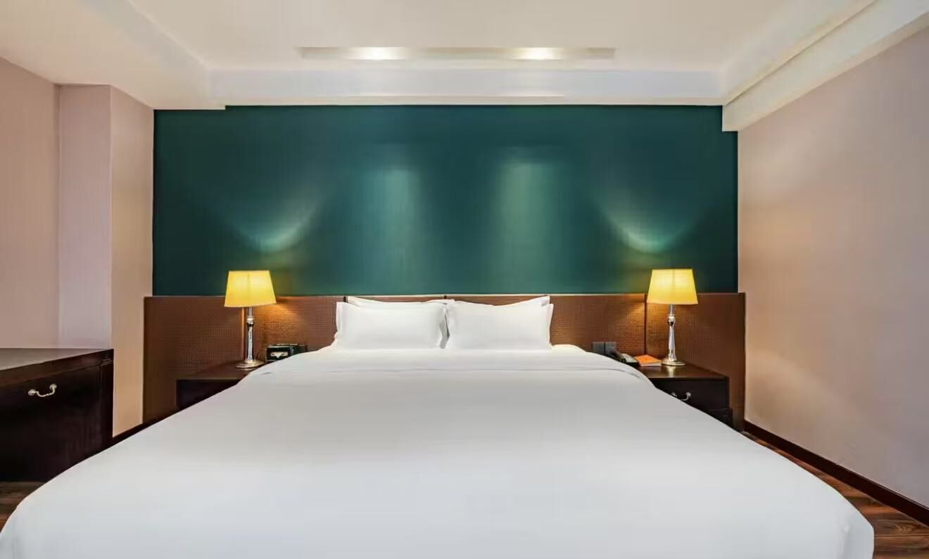 A hotel room with a large bed, two bedside tables with lamps, a dark green accent wall, and a wooden headboard.