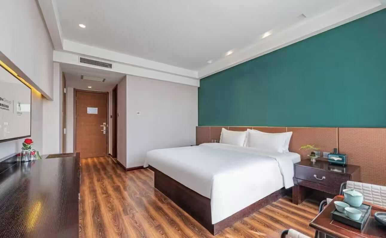 A hotel room with a large bed, a wooden floor, a desk with a chair, and a nightstand with a lamp.
