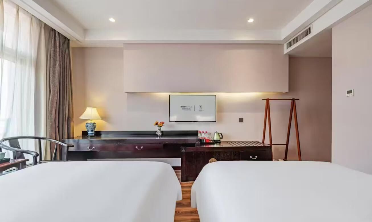 A hotel room with two beds, a desk with a lamp, a television, and a small table with a vase.