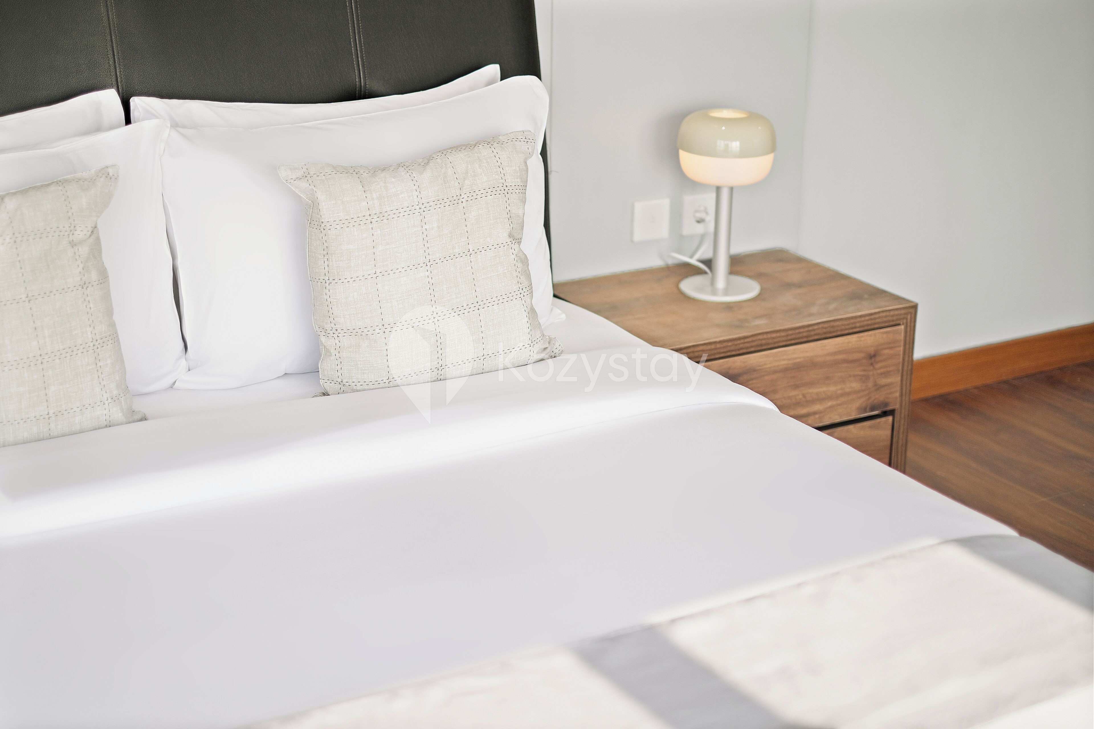 A neatly made bed with white linens and a headboard, a bedside table with a lamp, and a wooden nightstand.