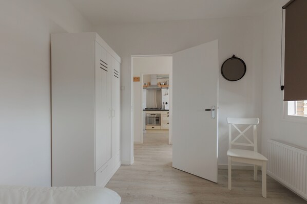 A room with a white wardrobe, a white chair, and a view into a kitchen area.