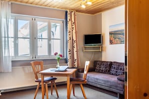 Une pièce avec une table en bois, deux chaises, un canapé, un téléviseur posé sur un meuble et une fenêtre avec des rideaux.