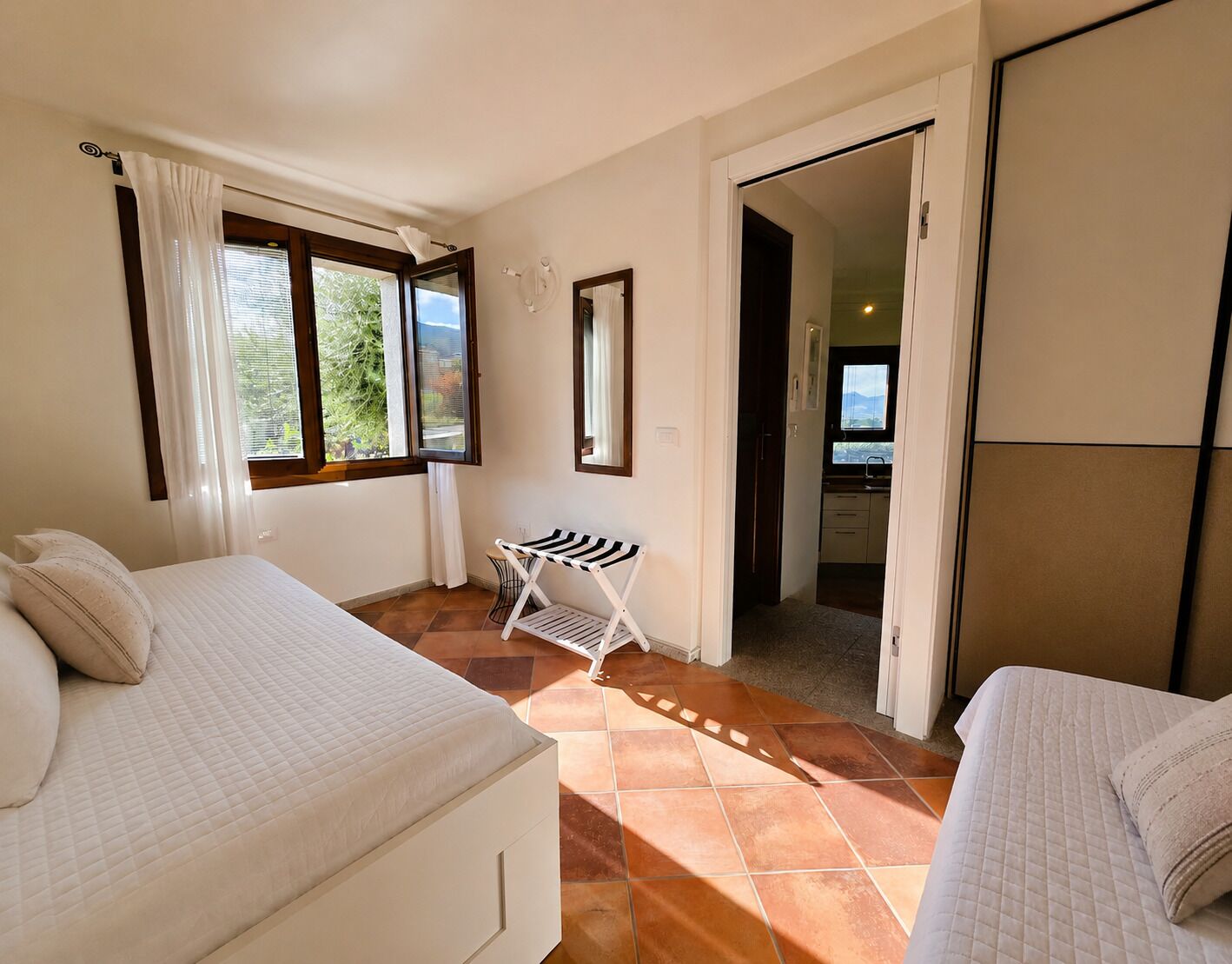 A bedroom with two beds, a window with curtains, a small table, and a view of the outside.