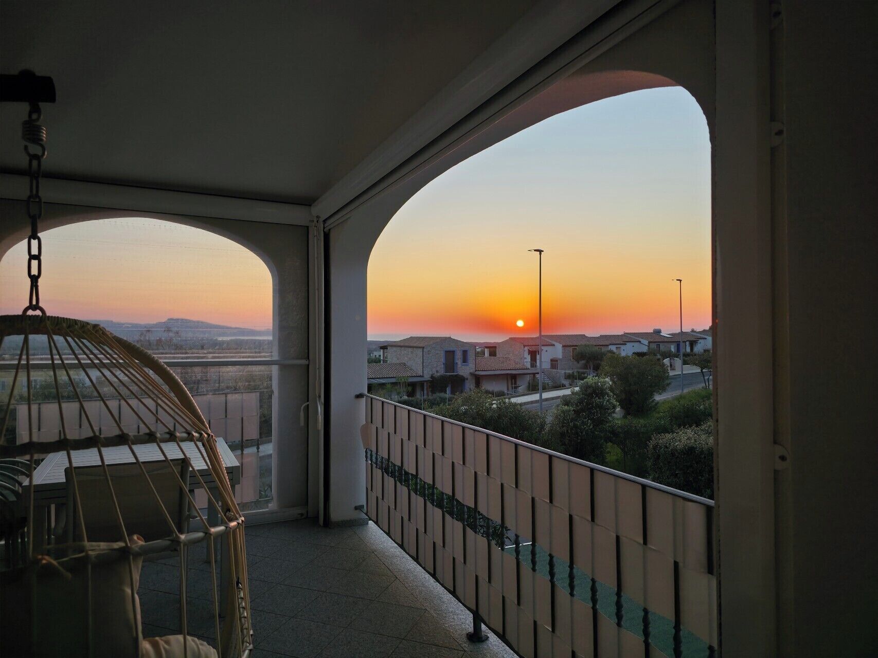 A balcony with a view of the sunset, a hanging chair, and a railing.