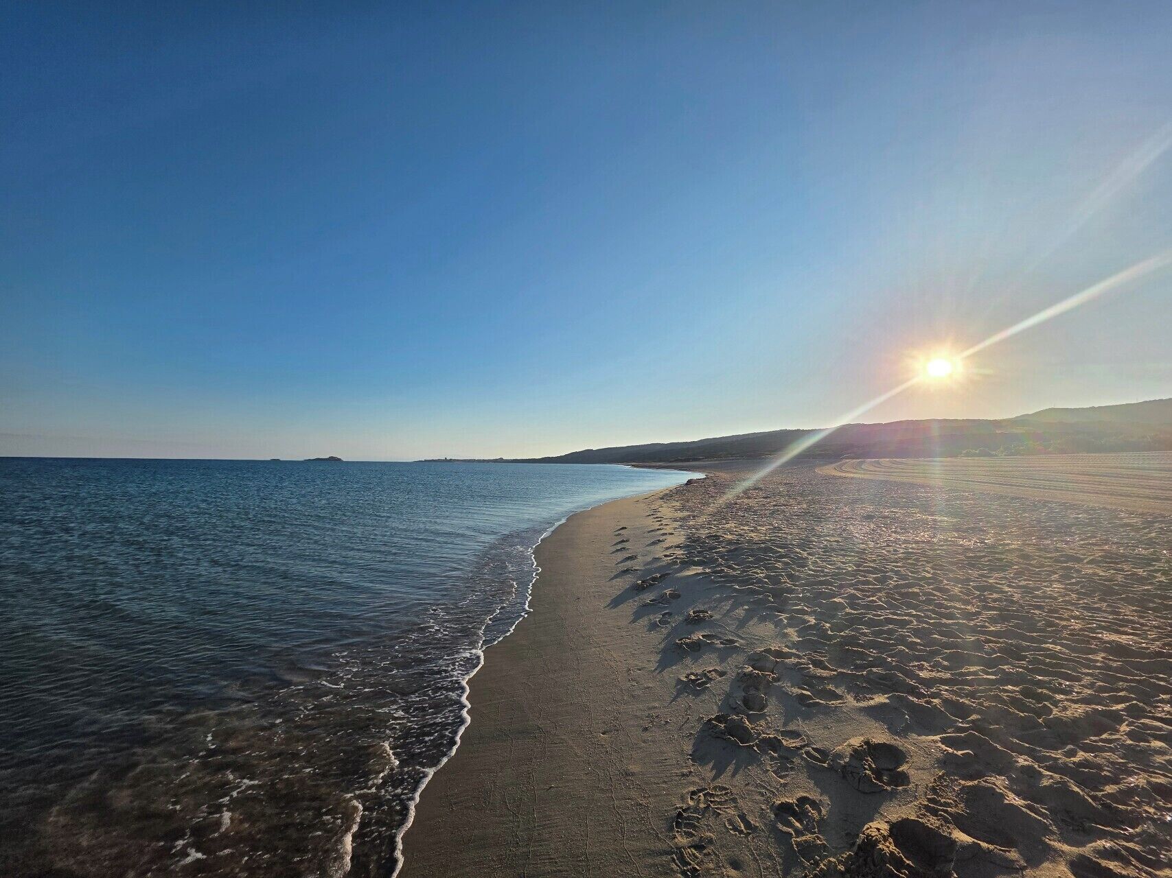 A beach with footprints leading to the water, a clear sky, and the sun setting or rising.