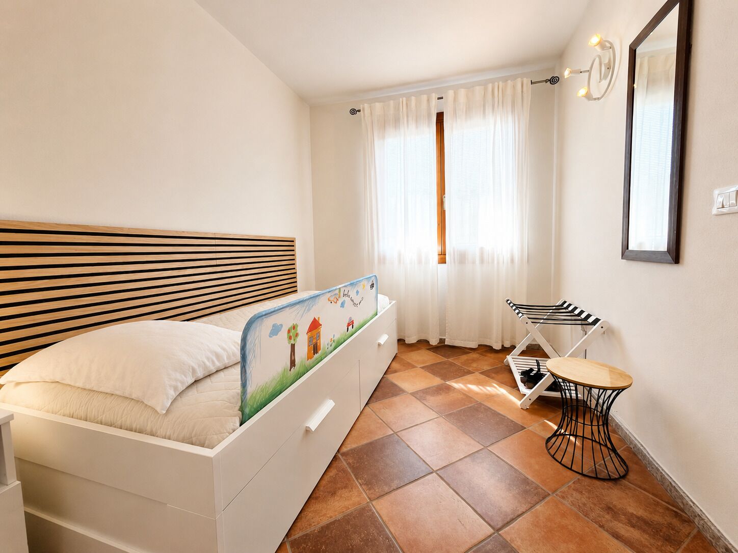 A bedroom with a bed, a small table, a chair, a window with curtains, and a mirror on the wall.