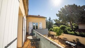 A small outdoor patio with a table and chairs, a door leading to a covered area, and a view of a tree and garden. - CosyFamily 1st floor-Pool-Wifi-Family-Plage (Pérols)
