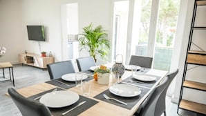 A modern dining area with a long wooden table set for four, surrounded by black chairs. A television is mounted on the wall, and a potted plant is visible in the background. - CosyFamily 1st floor-Pool-Wifi-Family-Plage (Pérols)
