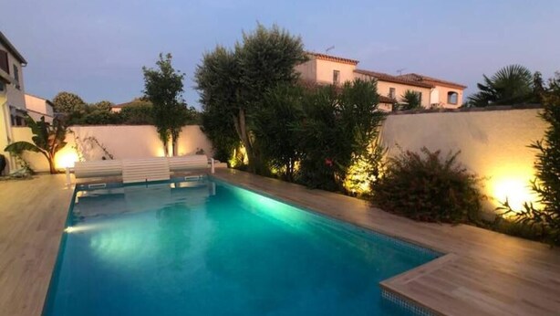 Une piscine rectangulaire entourée d’une terrasse en bois, avec en arrière-plan un paysage urbain composé d’immeubles et d’arbres.