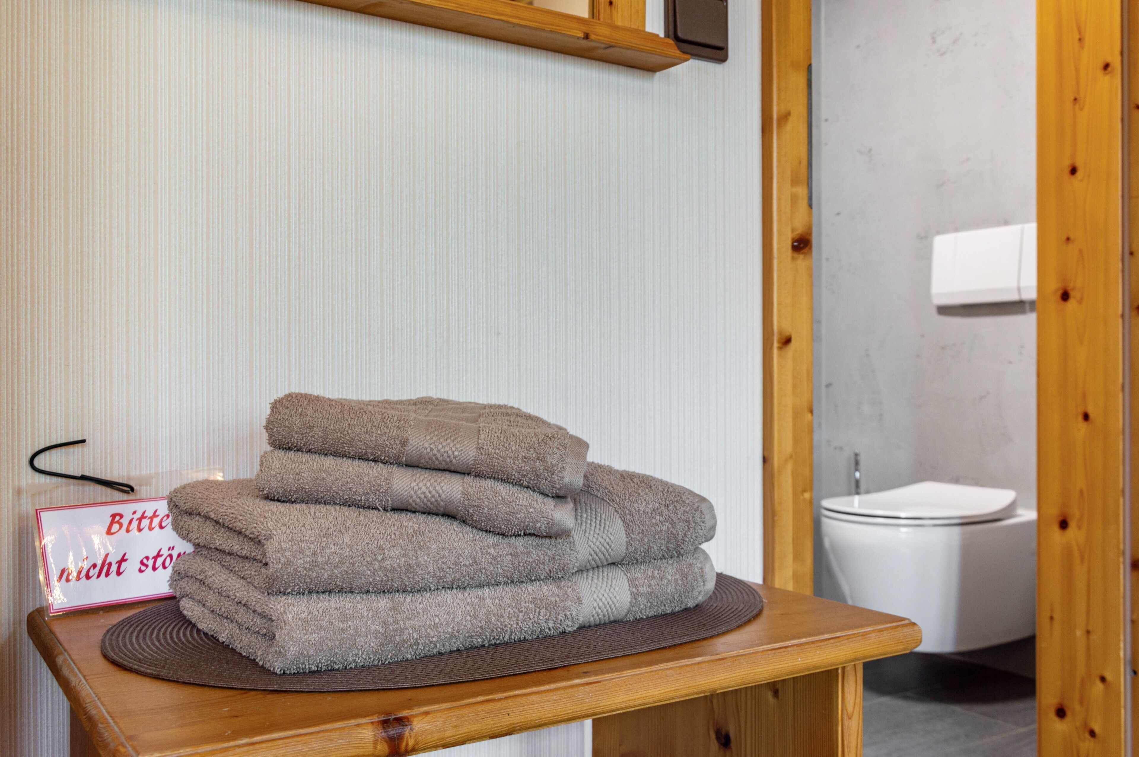 Een stapel handdoeken op een houten tafel met een bordje waarop staat 'Bitte nicht stören' in een badkamer met een toilet en een houten deur.