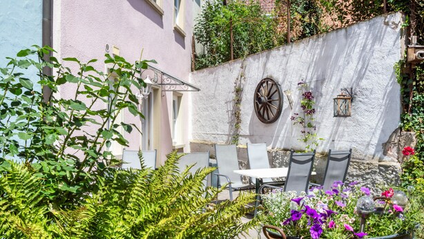 A patio area with outdoor seating, a white wall with decorative elements, and lush greenery. - Room 'Lutz - Zimmer 2' with Shared Terrace, Shared Garden and Wi-Fi (Iphofen)