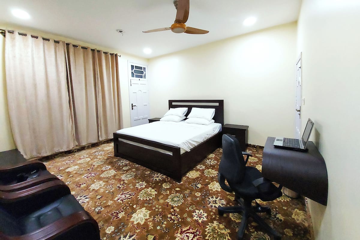 Une chambre d’hôtel avec un lit, un bureau avec un ordinateur portable, une chaise, un ventilateur de plafond et une moquette à motifs.