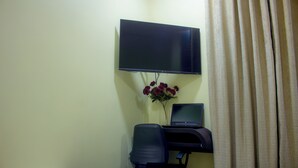 A room with a flat-screen TV mounted on the wall, a laptop on a desk, a chair, and a vase with flowers.