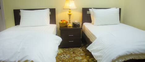 A hotel room with two single beds, a nightstand with a lamp, and a fruit basket.