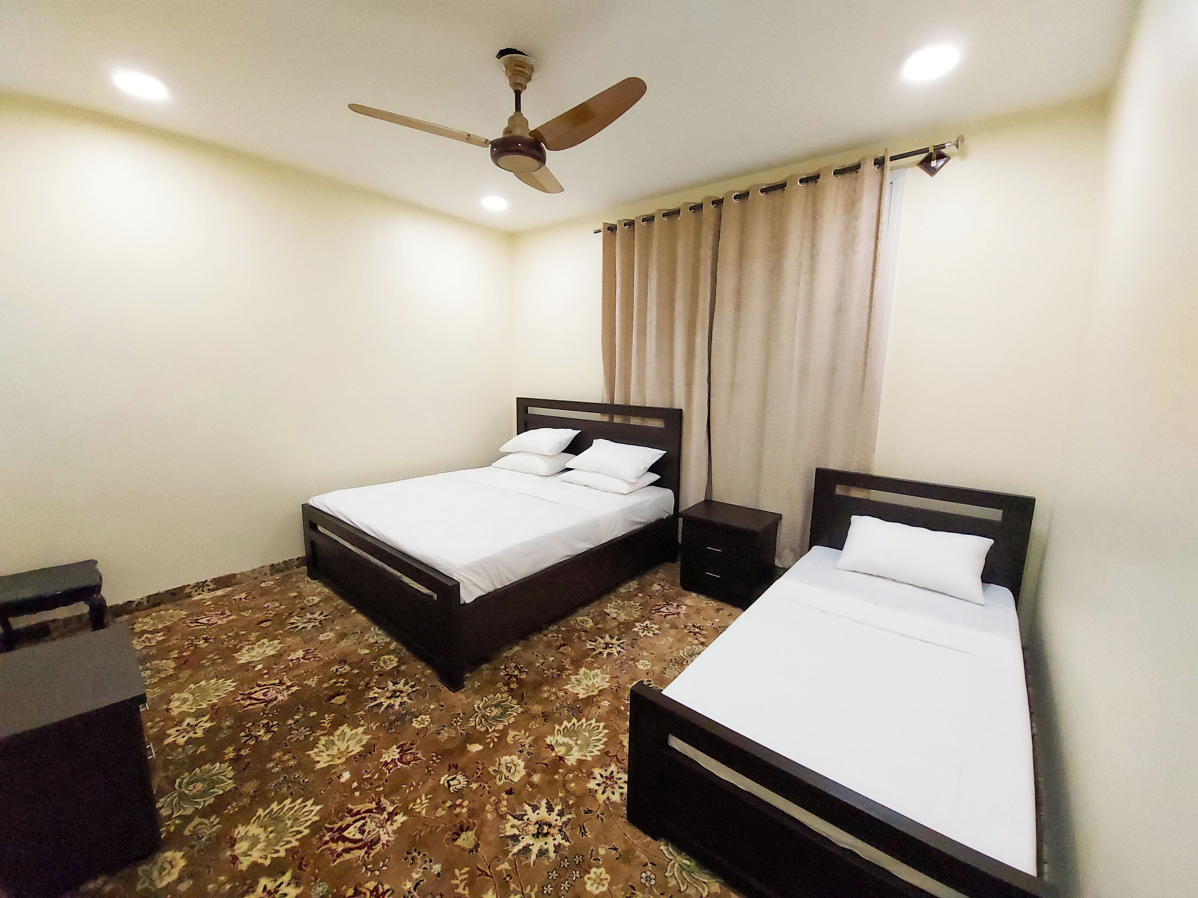A hotel room with two single beds, a ceiling fan, a nightstand, and a patterned carpet.