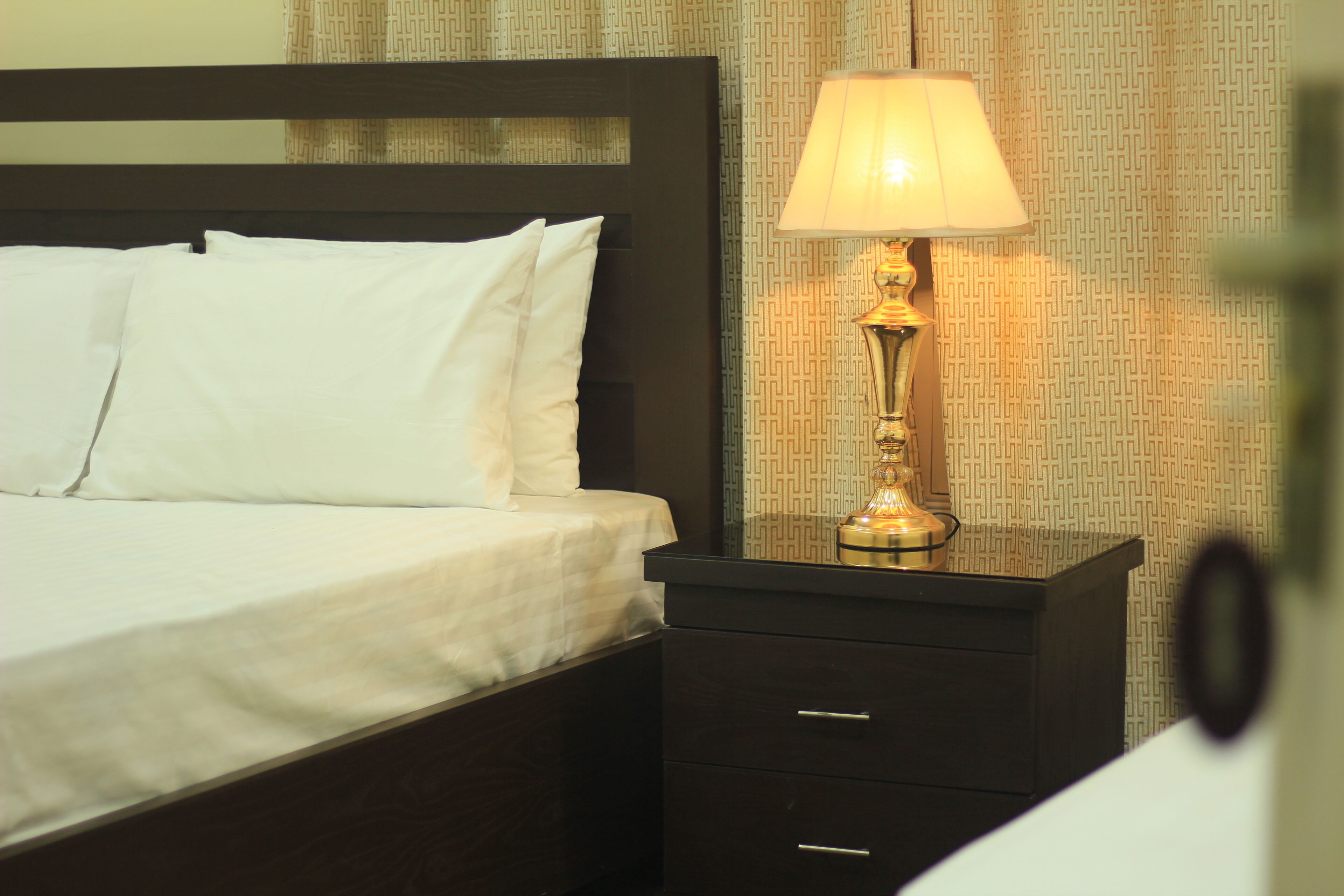 A neatly made bed with white linens and a dark wooden headboard. A bedside lamp with a gold base is on a matching dark wooden nightstand.