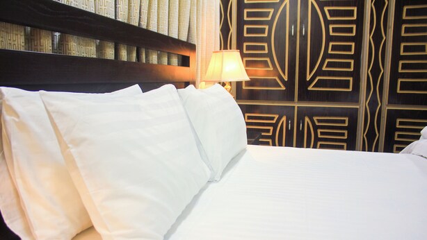 A neatly made bed with white linens and pillows, a bedside lamp, and a patterned wall in the background.