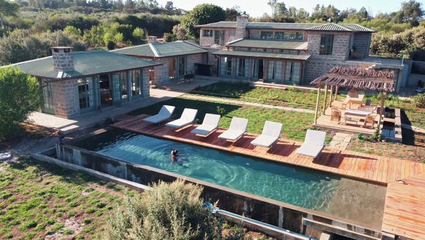 Una gran piscina con sillas de descanso blancas y una casa de piedra con techo verde.