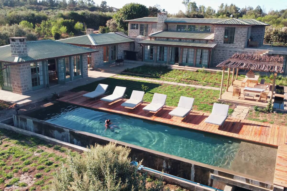 A large swimming pool with white lounge chairs and a stone house with a green roof.