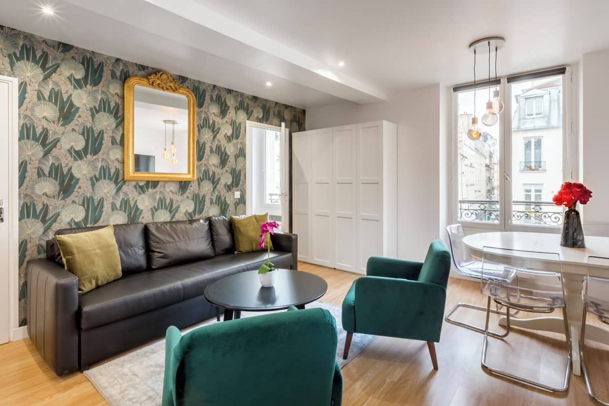 Una sala de estar moderna con un sofá de cuero negro, sillones verdes, una mesa redonda de centro y una mesa de comedor con sillas. Hay un espejo grande en la pared, una ventana con vistas a edificios y un jarrón con flores sobre la mesa de comedor.