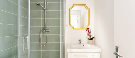 Casa de banho moderna com chuveiro em cabine de vidro, lavatório branco com espelho emoldurado a dourado e uma pequena orquídea em vaso.