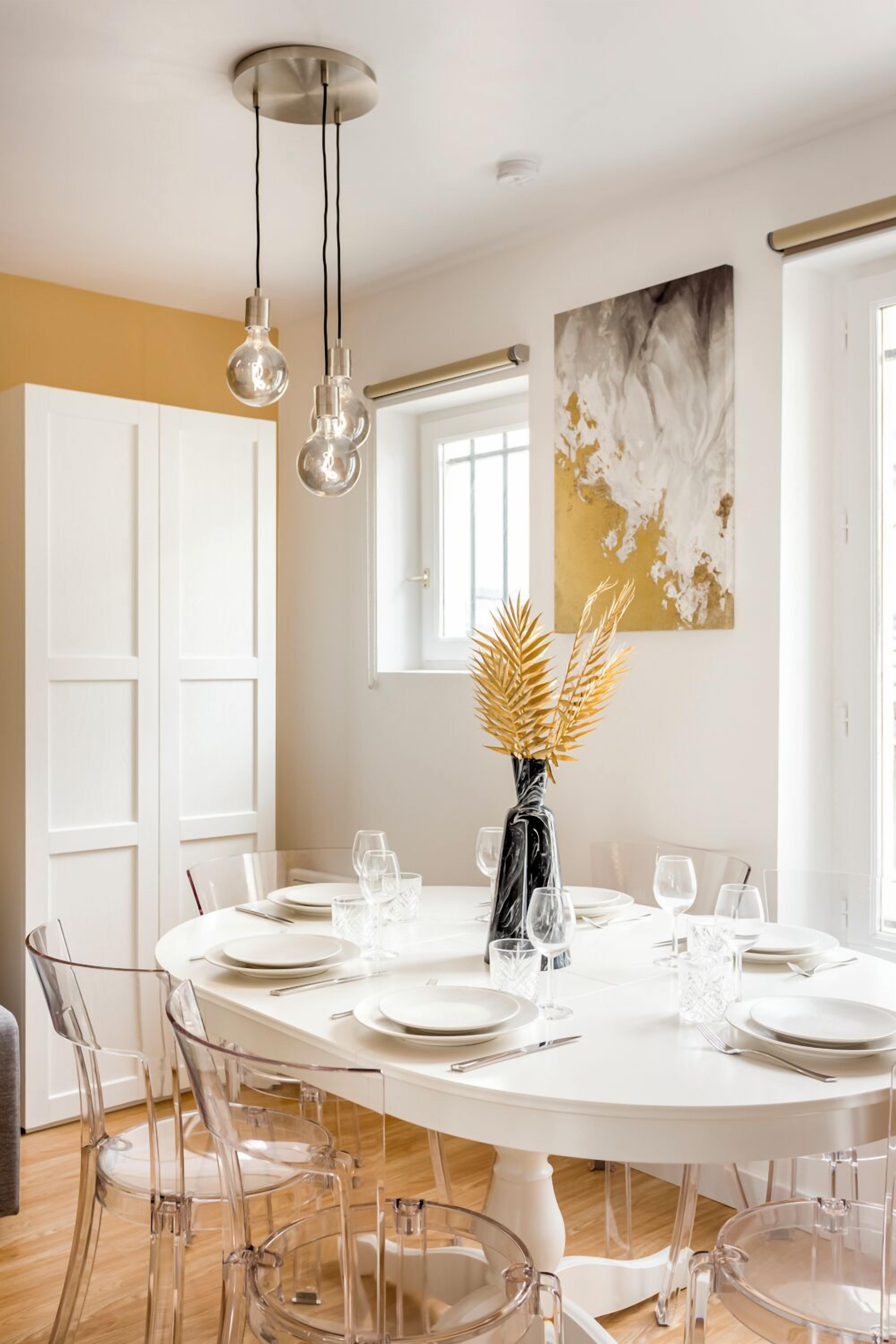 A dining area with a round table set for six, clear chairs, and a vase with dried plants.