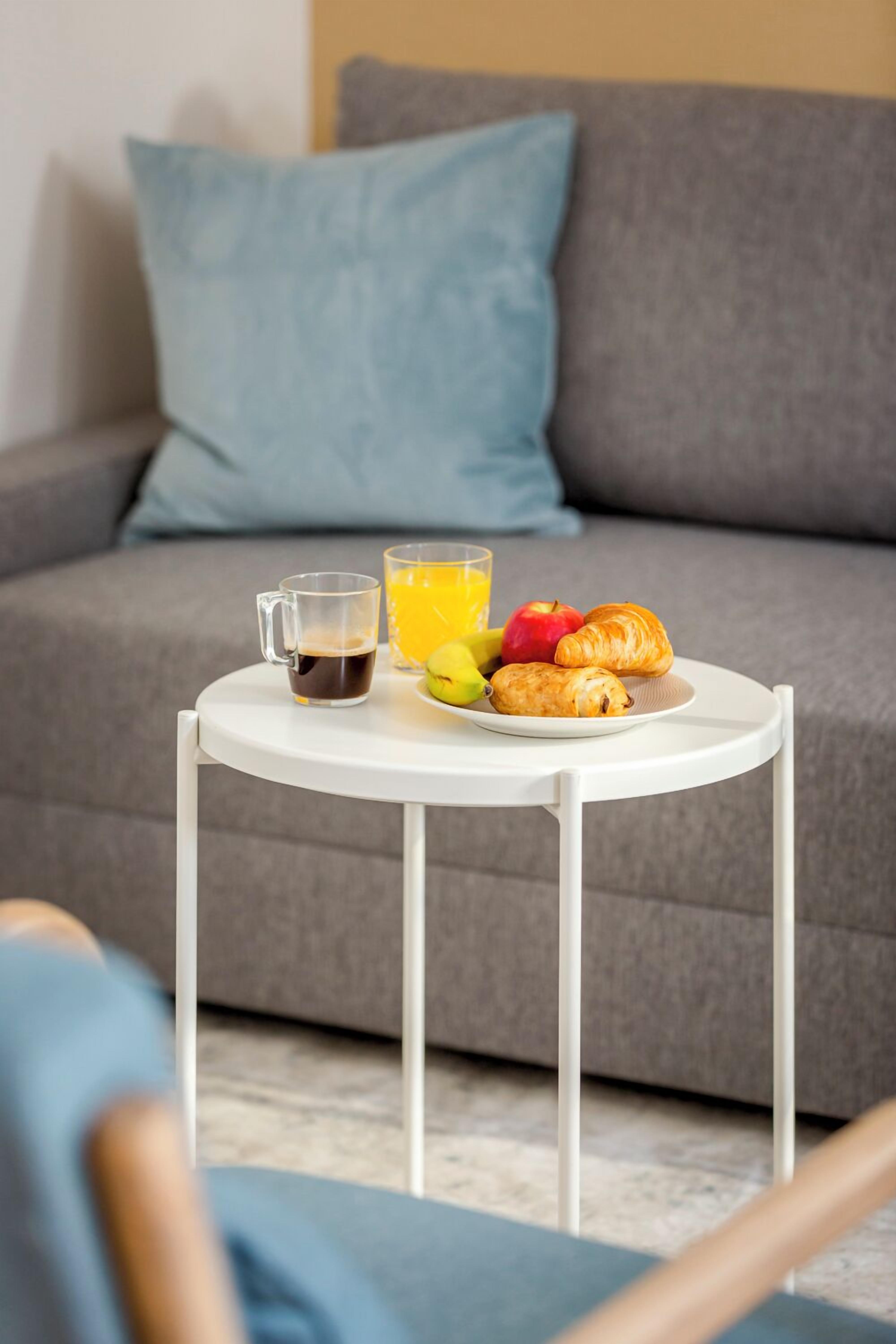 A breakfast setup on a small white table with a beverage, fruit, and pastries in front of a grey sofa with blue cushions.