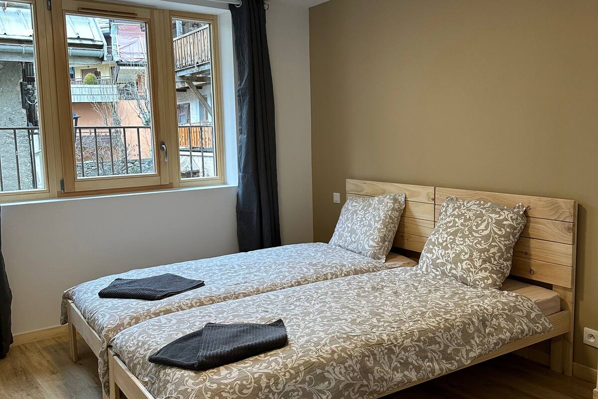 Un lit double avec une couvre-lit à motifs, une tête de lit en bois et une fenêtre donnant sur les bâtiments environnants.