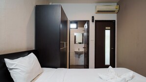 A hotel room with a bed, a white pillow, a neatly folded towel, a dark wardrobe, a bathroom with a sink and mirror, and an air conditioning unit.