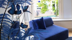 Una habitación con un sofá azul, un ventanal con vistas al verde exterior y una planta decorativa en primer plano.