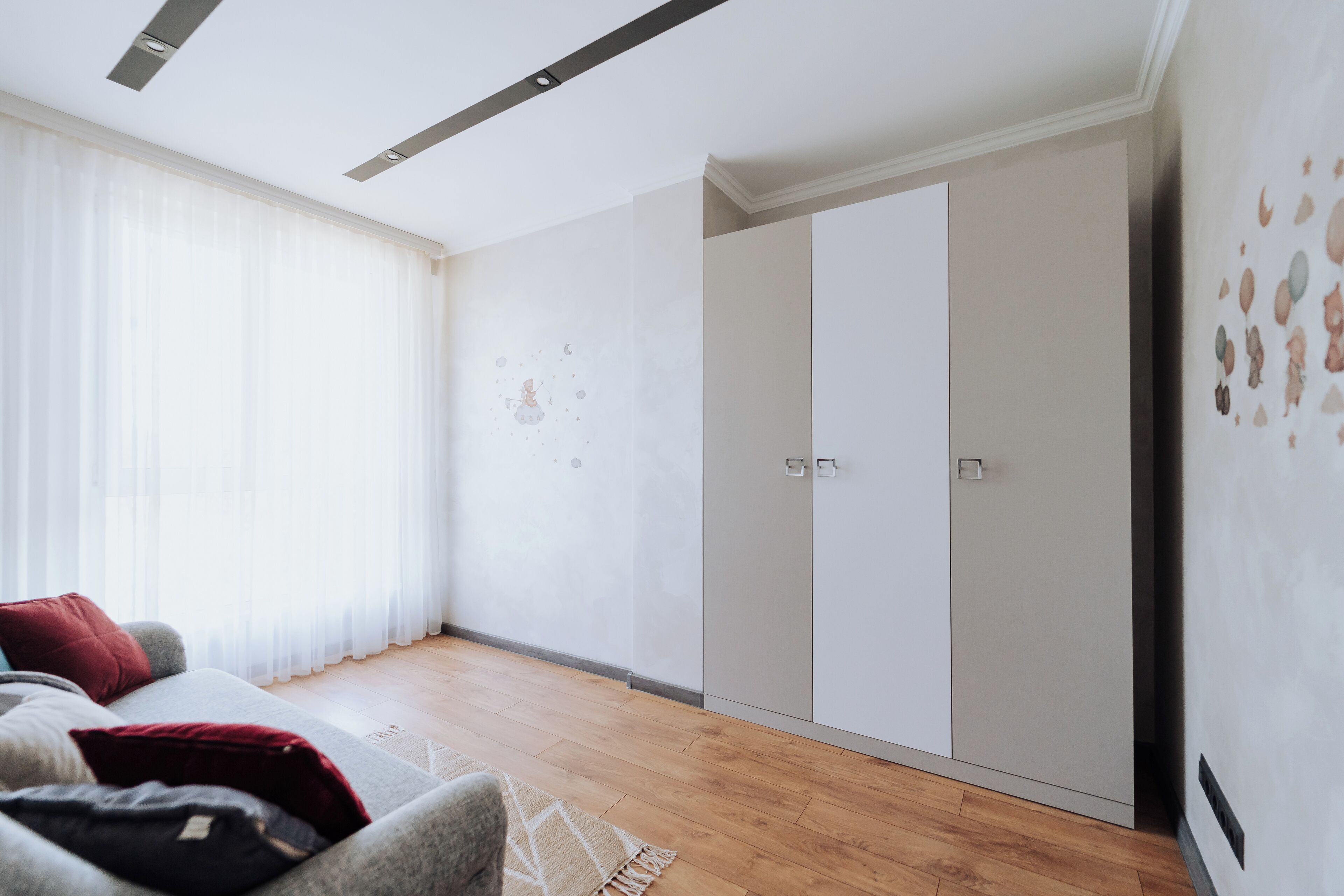 A modern living room with a grey sofa, a red pillow, and a wooden floor. There is a large wardrobe with two doors and a wall with a decorative pattern.