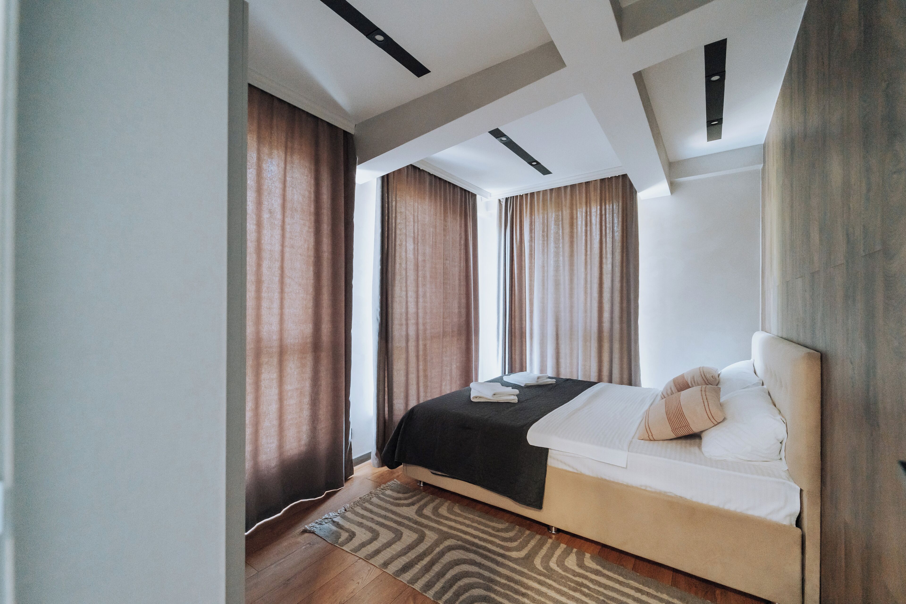 A modern hotel room with a bed, curtains, and a ceiling with recessed lighting.
