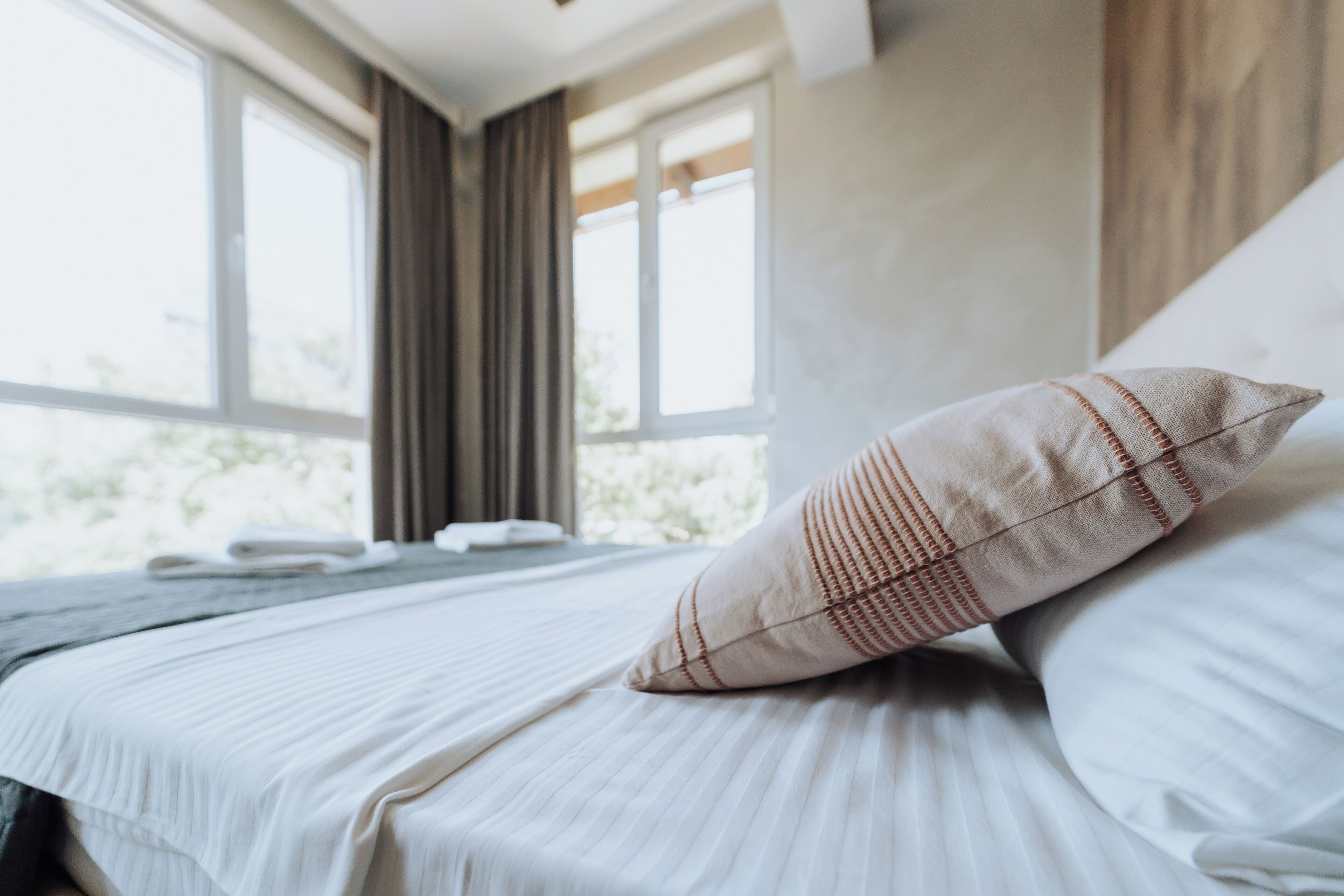 A neatly made bed with white linens and a striped pillow, large windows with curtains, and a view of greenery outside.