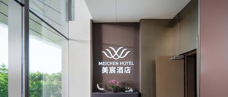 A reception area with a sign for Meichen Hotel, a reception desk, potted plants, and a view of greenery outside.