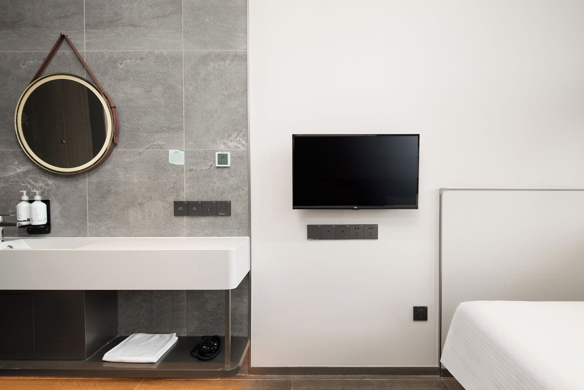 Habitación de hotel moderna con un espejo redondo grande, un televisor de pantalla plana en la pared, un lavamanos blanco con artículos de aseo y una cama con edredón blanco.