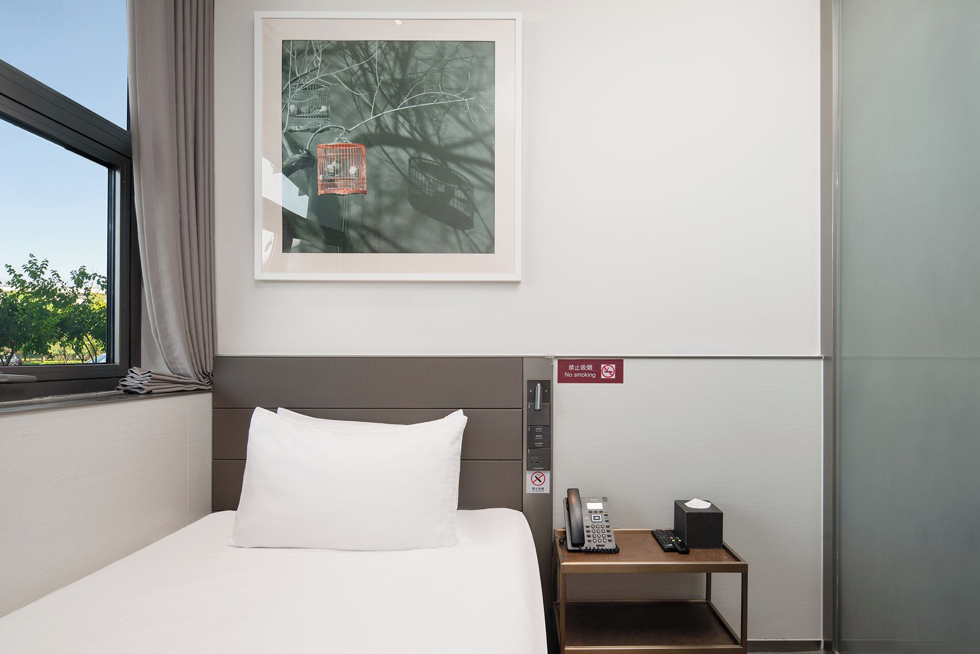 A hotel room with a bed, a nightstand with a phone and tissues, a framed picture of birds, and a no-smoking sign.