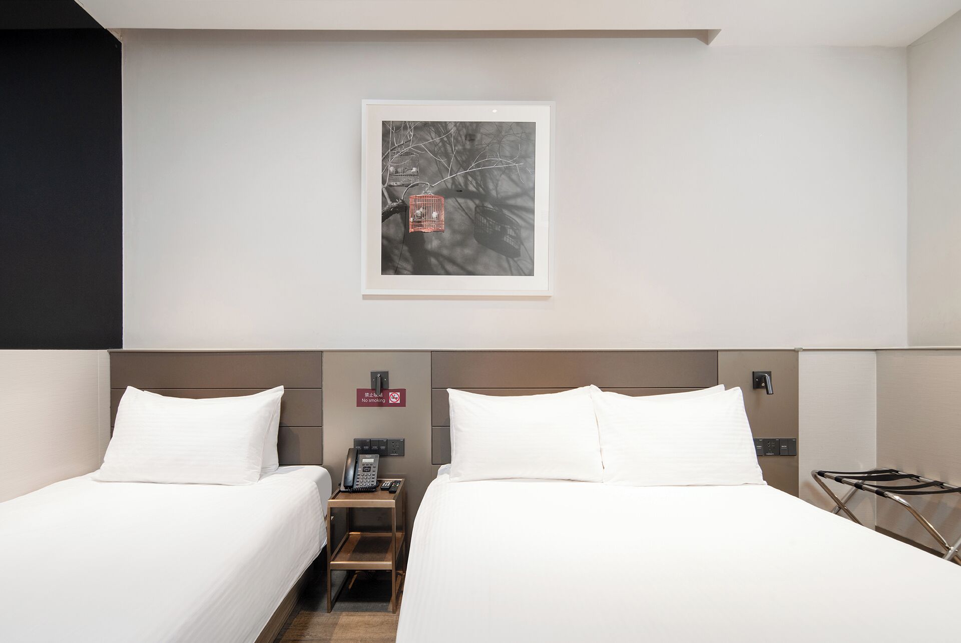 Une chambre d’hôtel avec deux lits, un tableau encadré au mur, un téléphone sur la table de chevet et un panneau indiquant qu’il est interdit de fumer.