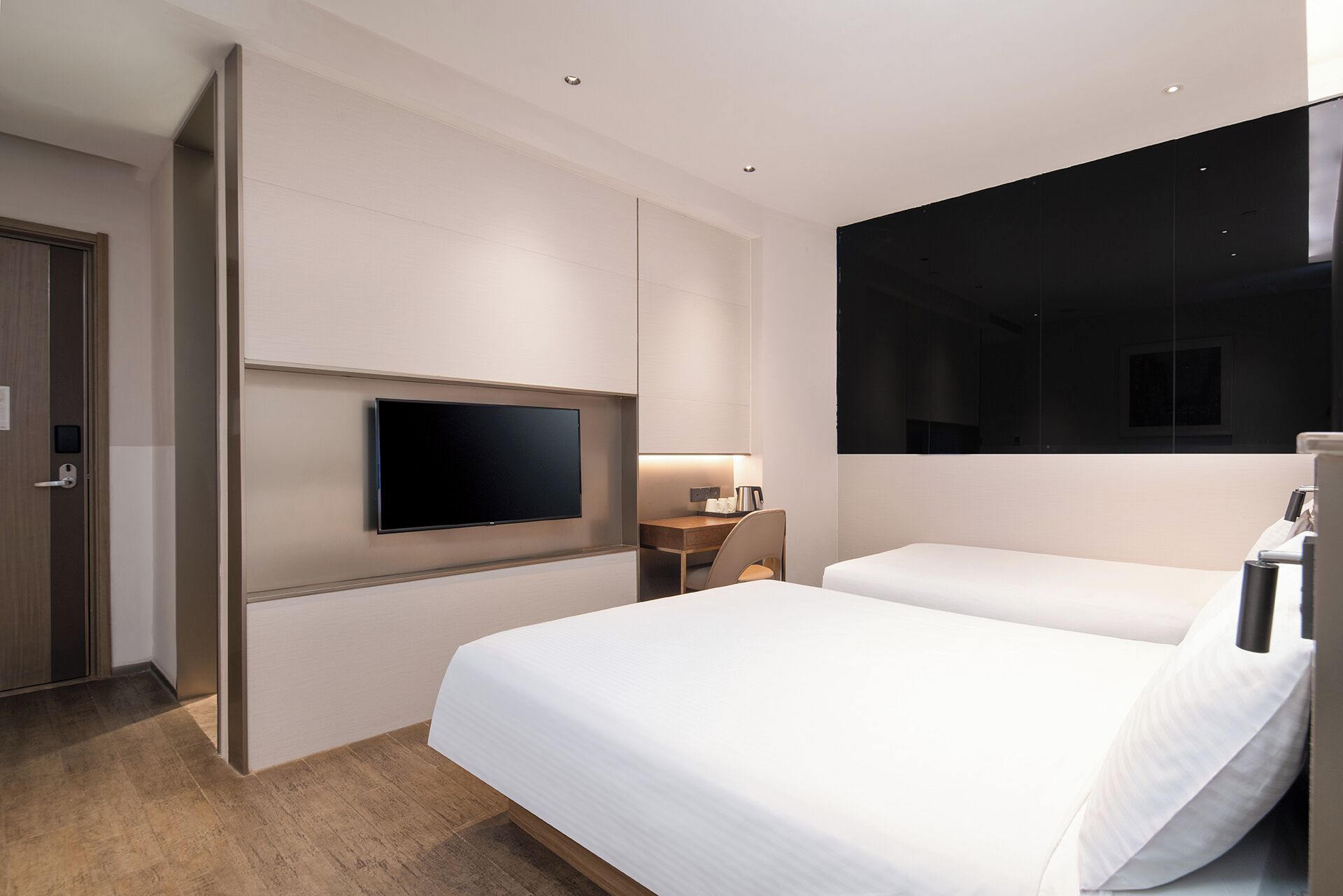 A modern hotel room with a large bed, a flat-screen TV mounted on the wall, a desk with a chair, and a large window.