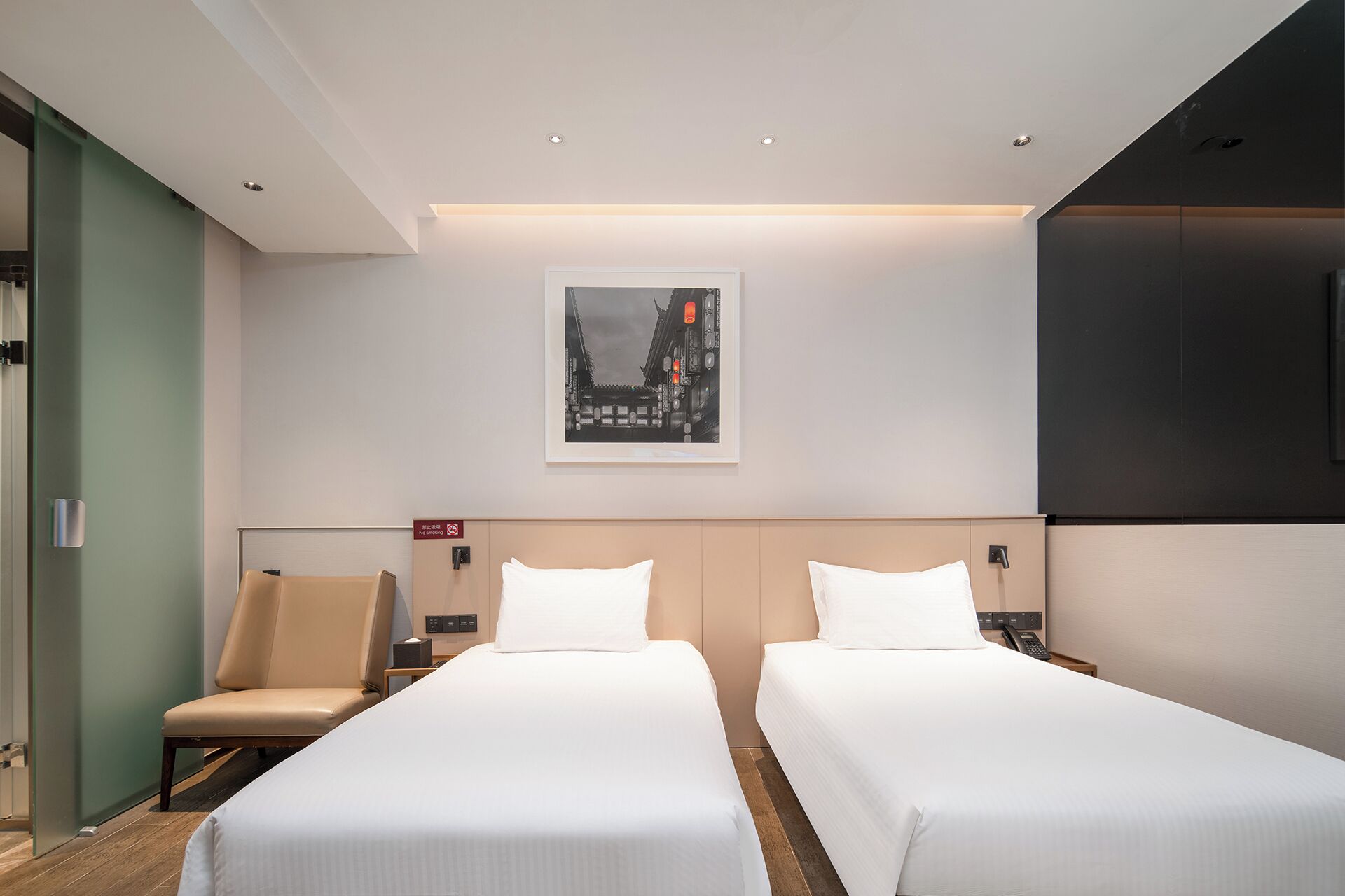 A hotel room with two beds, a chair, a nightstand, a wall-mounted TV, and a framed picture on the wall.