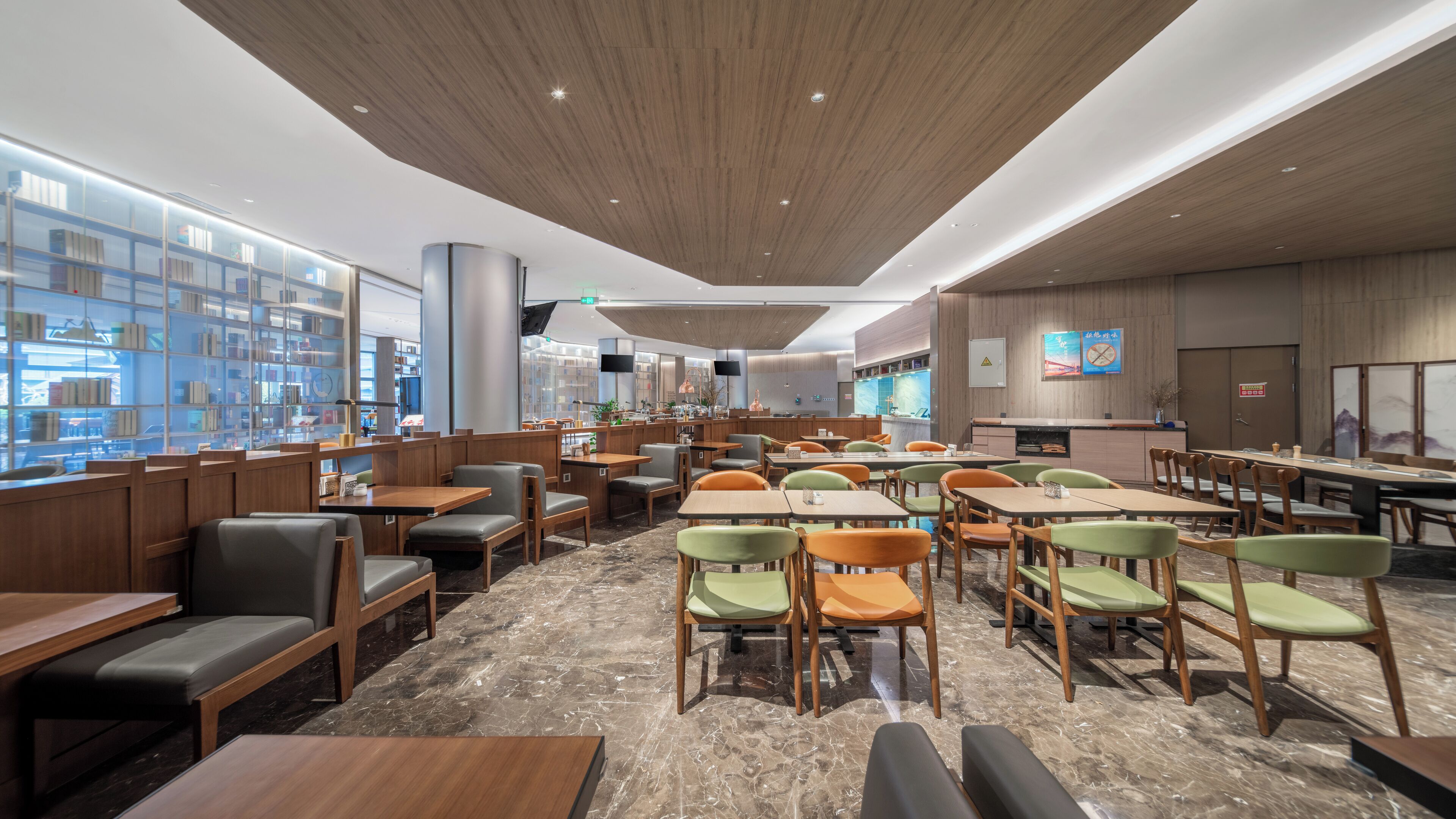 A modern restaurant with wooden booths, green and orange chairs, marble flooring, and a large bookshelf area.