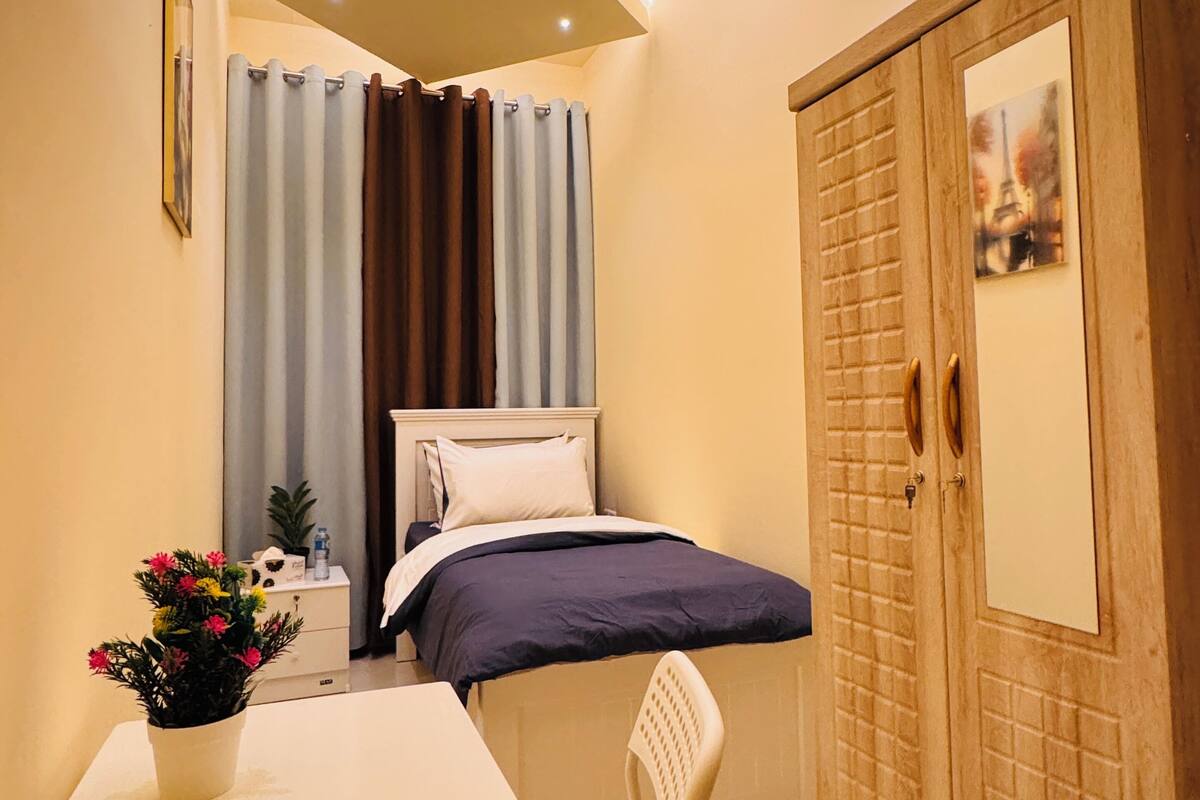 A small, well-lit room with a bed, a wooden wardrobe, a table with a chair, and a potted plant.