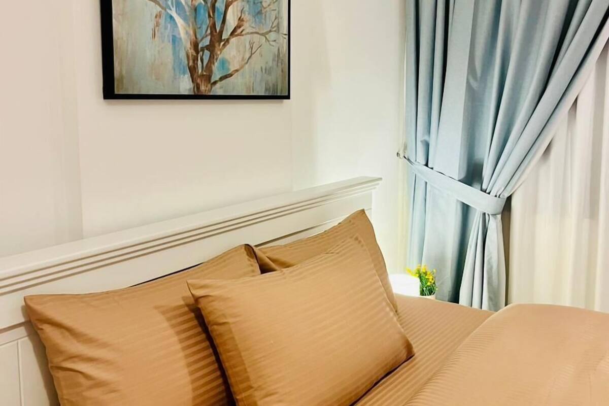 A hotel room with a bed, beige bedding, a painting of a tree, and light blue curtains.
