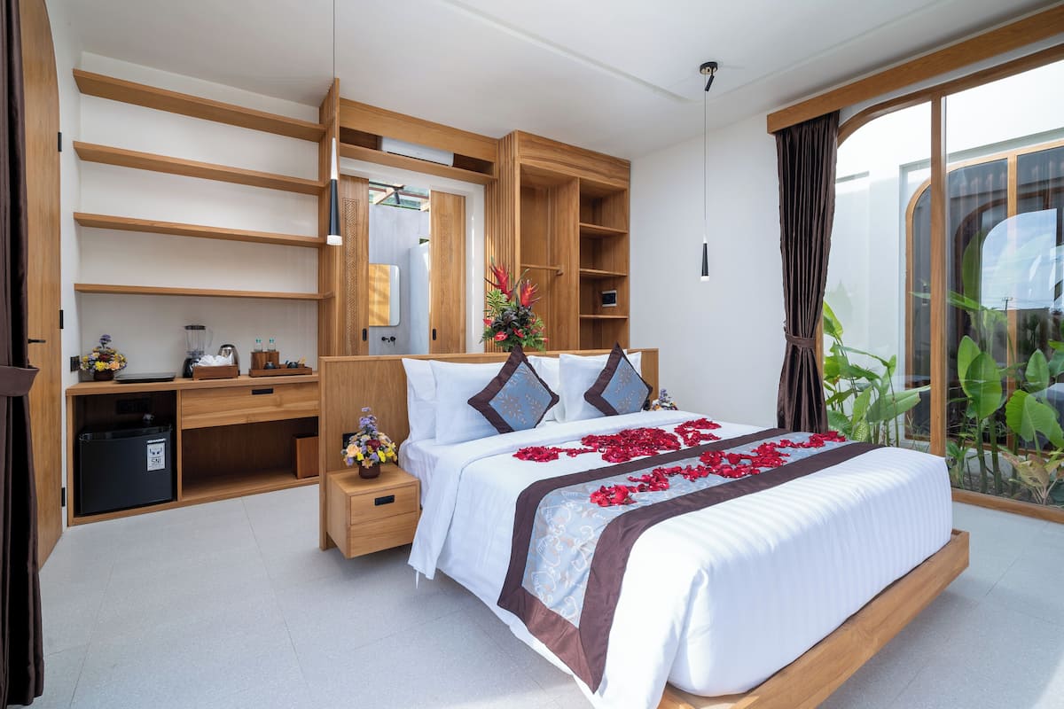 A modern bedroom with a large bed, wooden built-in shelving, a small desk, and a view of greenery through a large window.