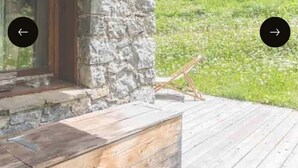 Una terraza de madera con paredes de piedra, un ventanal y vista a una colina cubierta de césped.