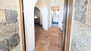 A hallway with stone walls and a tiled floor leading to a bedroom with a bed and a nightstand. - Dammuso - Cala Cinque Denti - AFFITTI\'CA (Pantelleria)
