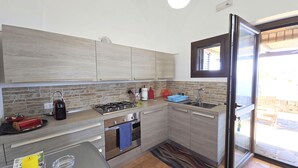 A modern kitchen with a gas stove, oven, and sink. There is a window with blinds, a door leading to an outdoor area, and a patterned rug on the floor. - Dammuso - Cala Cinque Denti - AFFITTI\'CA (Pantelleria)
