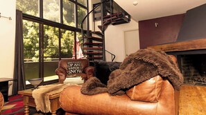 A cozy living room with a leather sofa, a fireplace, and a large window with a view of trees. - Bernti's Mountain Inn (Thredbo)
