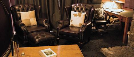 A room with two leather armchairs, a wooden table with a book and salt shakers, a lamp, and a stone accent wall.