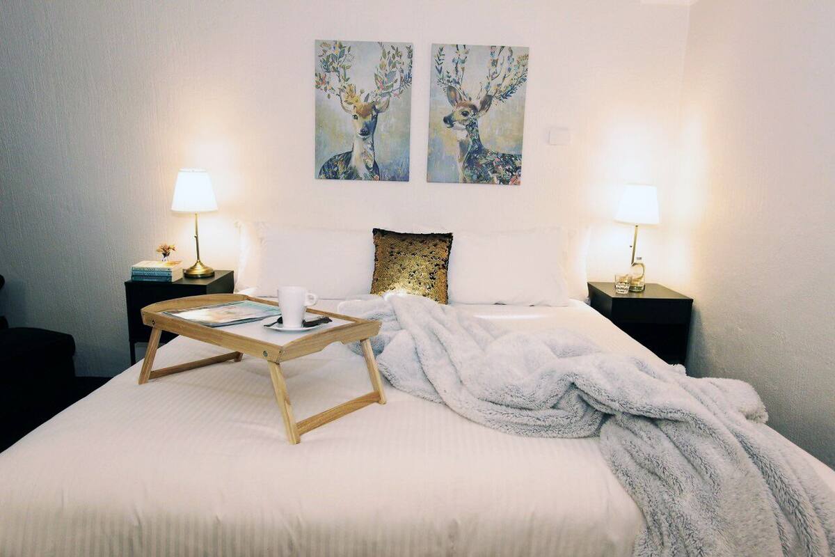 A neatly made bed with a white blanket, two bedside lamps, a small table with a cup and a book, and two wall-mounted paintings of deer.